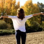 Una chica en plena naturaleza. Está de pie y de espaldas, con los brazos abiertos de par en par respirando aire puro para así liberarse de los compuestos orgánicos volátiles que se pueden respirar en los interiores de las casas.