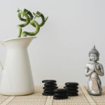 Un jarro con dos plantas bambúes, 3 apilamientos de piedras zen y una estatua pequeña de buda con las manos en postura de gratitud. Una escena zen y de dar gracias.