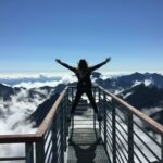 Mujer joven de espaldas en un mirador alto en una montaña. Está asomada con los brazos abiertos en alto, símbolo de éxito.