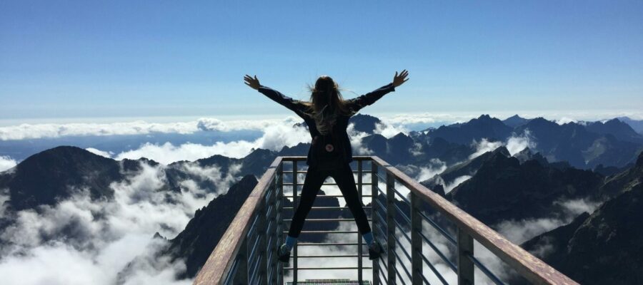 Mujer joven de espaldas en un mirador alto en una montaña. Está asomada con los brazos abiertos en alto, símbolo de éxito.