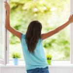Mujer joven de espaldas abriendo una ventana para ventilar la casa con aire del exterior.