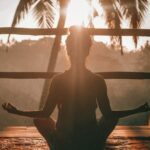 Mujer de espaldas, a contraluz, en una terraza de madera practicando la meditación zen. Ambiente calmado y de atardecer relajado.