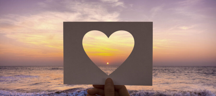 Puesta de sol en una playa enmarcada por una figura de un corazón recortado en un papel. Colores suaves y ambiente muy romántico.