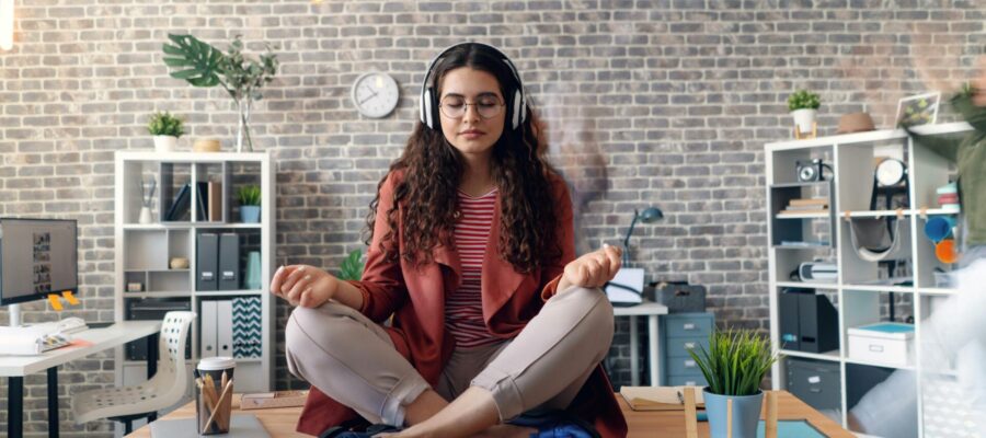 Chica joven sentada sobre la mesa de una oficina y meditando con los ojos cerrados y con los cascos escuchando la meditación. Oficina zen y enfocada a la productividad.