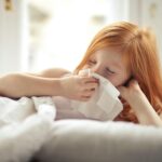 Niña pelirroja en la cama sonándose la nariz. Tiene alguna alergia provocada por algún alérgeno que está en su casa.